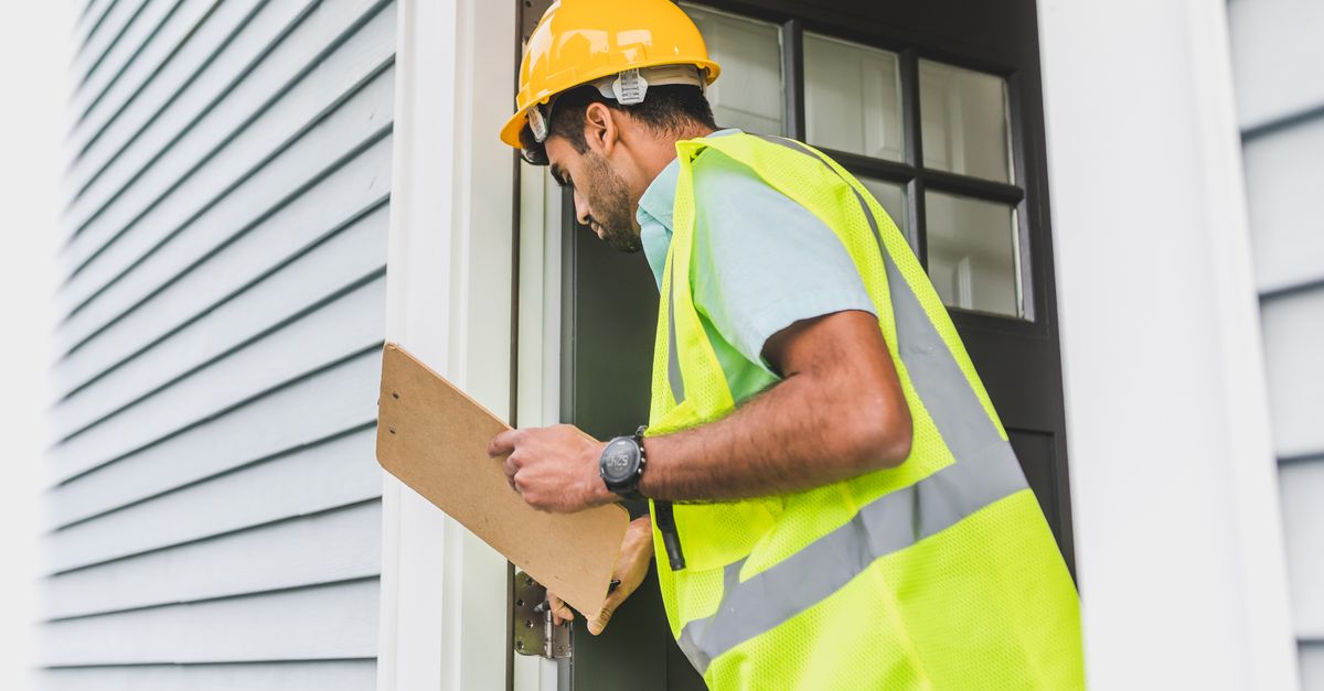 A male inspector in a hard hat conducts a property inspection outdoors with a clipboard.