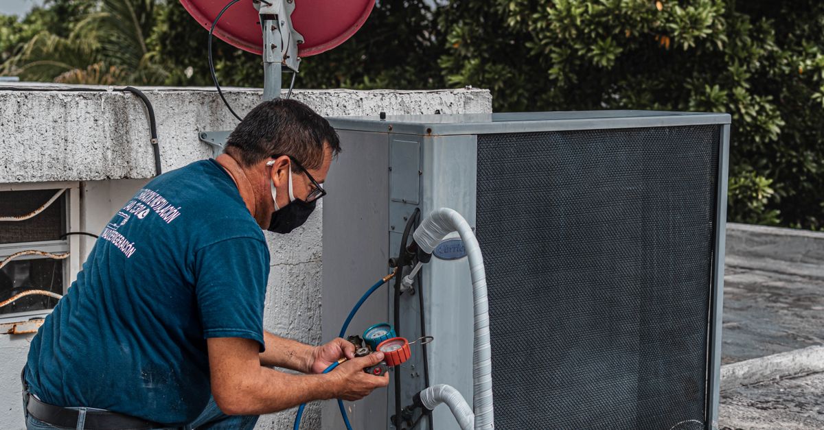 Technician repairing an air conditioner unit outdoors, wearing a facemask and using a manifold gauge.